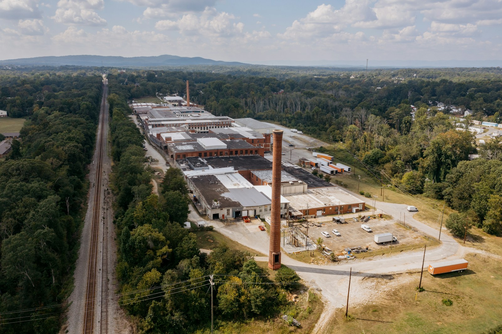 Real aerial photograph of the Taylors Mill industrial complex, showing red-brick factory buildings, tall smokestack, adjacent industrial land and tree-lined surroundings in South Carolina
